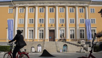 Keltainen suuri rakennus, johon johtaa leveä portaikko. Edessä kaksi pyöräilijää.