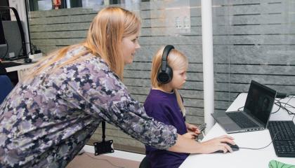 Pieni vaaleahiuksinen tyttö istuu tietokoneen äärellä kuulokkeet korvillaan. Hänen takanaan on tytön äiti, joka pitelee pöydällä olevaa tietokoneen hiirtä.