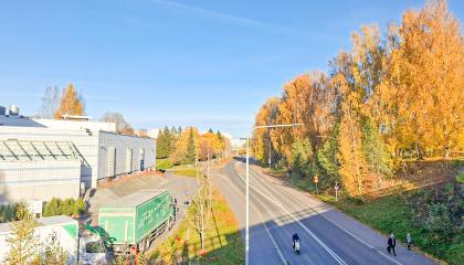 Autotie, pyörä- ja jalankulkuväylät ja kulkijoita syysväreissä olevien puiden ympäröimän, vasemmalla rakennus.