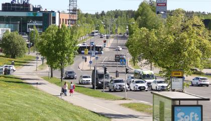 Vihreää nurmikkoa ja puita, jalankulkijoita ja autoja pitkällä tiellä.