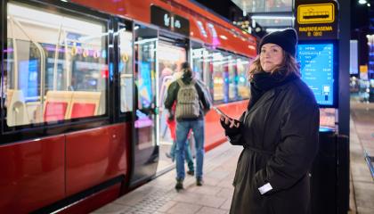 Henkilö seisoo ratikkapysäkillä kädessään älypuhelin. Ratikka on pysähtynyt ja matkustajia on menossa sen sisään.