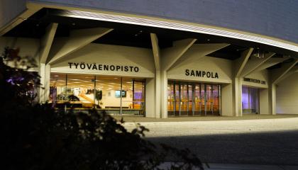 Sampola-rakennuksen pääsisäänkäynti iltavalaistuksessa. Lasiseinien läpi näkyy valaistu aula.