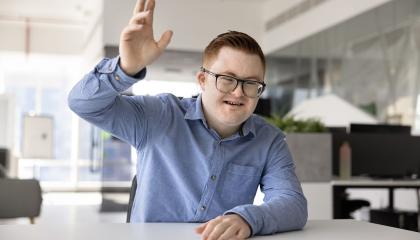 Kauluspaitaan pukeutunut henkilö, jolla on Downin oireyhtymä, nostaa toista kättään.