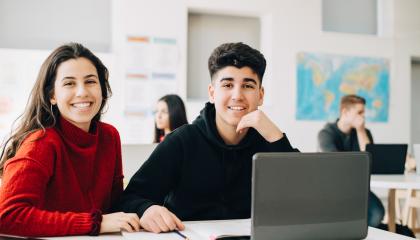 kuvassa kaksi henkilöä istuu luokkahuoneessa tietokoneen ääressä.