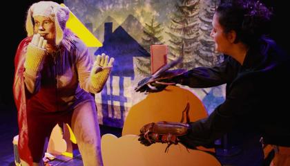 Koiran roolipuvussa oleva ihminen pitää toista &quot;tassua&quot; suun edessä ja hymyilee. Taustalla kuusia sekä varjokuva talosta.