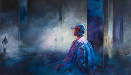 Nuori lippispäinen mies seisoo hämyisessä, tyhjässä tehdashallissa. 