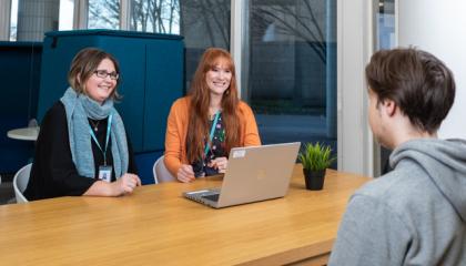 Kuvassa kolme henkilöä. Kaksi istuu pöydän toisella puolella kasvot kuvaajaan päin ja yksi selin. Kaikki hymyilevät. Heidän välissään on tietokone ja menossa on jonkinlainen ohjaustilanne.