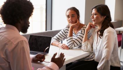 Teini-ikäinen nuori ja äiti istuvat pöydän ääressä. Pöydän toisella puolella istuu opettaja tietokoneen kanssa.