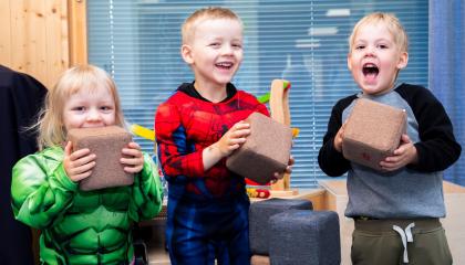 Kolme pientä lasta katsoo kameraan nauraen pitäen pieniä ruskeita pehmokuutioita kädessään.