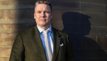 Henkilö seisoo tummassa puvussa tumman seinän edessä. Vieressä näkyy tumma varjo.