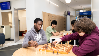 Neljä henkilöä pelaa shakkia, kaksi edessä ja kaksi takana pöydällä olevalla shakkilaudalla.