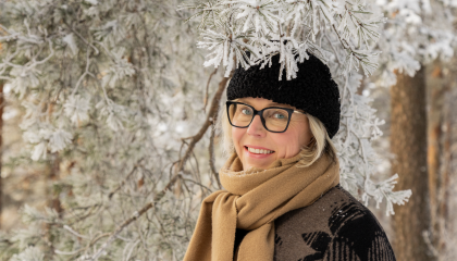 Henkilö hymyilee talvivaatteissaan lumisen puun alla.