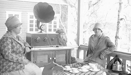 Mustavalkoisessa valokuvassa kaksi naista istuu ulkona kuuntelemassa vanhanaikaista radiota.