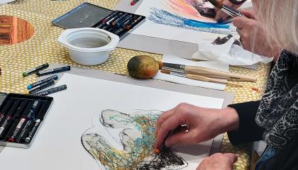 Harmaahiuksinen nainen maalaa värikkäillä liiduilla abstraktia teosta paperille.