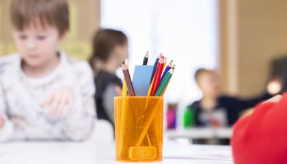 Kynäkuppi on pöydällä etualalla ja taustalla pieniä koululaisia.