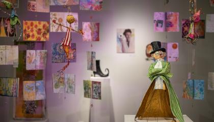 Kuva galleriasta, jossa etualalla lierihattupäinen veistos ja taka-alalla on katosta roikkuvia värikkäitä teoksia. 