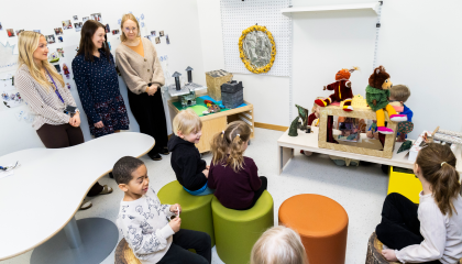 Lapset esiintyvät edessä ja toiset katsovat istuen. Päiväkodin työntekijät seuraavat tilannetta seisoen.