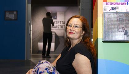 Mustaan lyhythihaiseen puseroon pukeutunut punahiuksinen nainen nojaa seinään. Taustalla erottuu museon näyttelyvitriini.