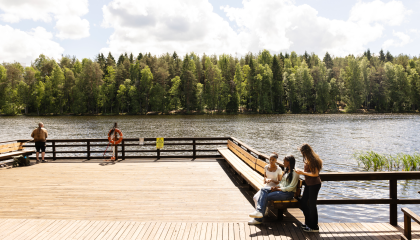 Yksi mies kalastamassa ja kolme nuorta tyttöä istumassa Ahvenisjärven uudistetulla laiturilla poutaisena kesäpäivänä. 