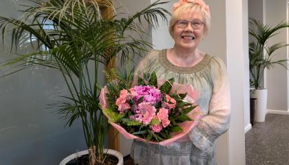 Vaaleahiuksinen hymyilevä Aija Karttunen seisoo viherkasvin vieressä. Hän pitelee käsissään suurta kukkakimppua, jossa on vaaleanpunaisia kukkia.