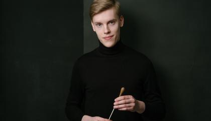Kapellimestari Kristian Sallinen mustassa poolossa, tahtipuikko kädessään mustaa taustaa vasten.