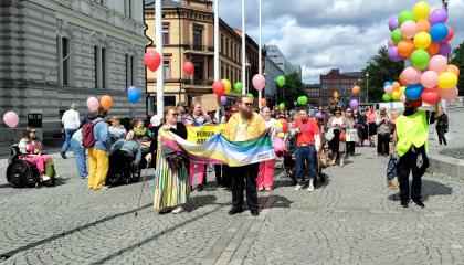 Vammaisten kulkue kävelee Keskustorilla. Etummaiset pitelevät käsissään pride-lippua. Monilla on värikkäitä ilmapalloja. Osallistujissa näkyy muun muassa pyörätuolilla kulkevia.