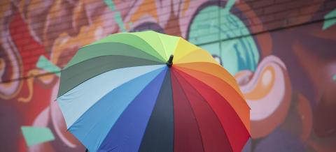 Ihminen seisoo monivärisen sateenvarjon alla suojassa.