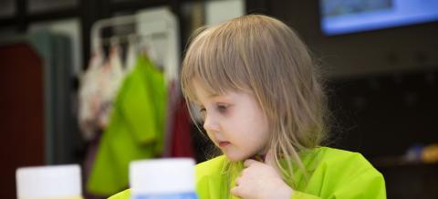 Takaa-alalla lapsi vihreässä essussa, edessä keltainen ja sininen iso maalipurkki.