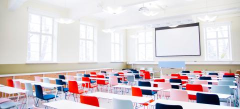 Valoisassa luentosalissa on riveissä valkoisia pöytiä ja värikkäitä, velkänojallisia tuoleja. Salin takaseinällä on valkokangas.