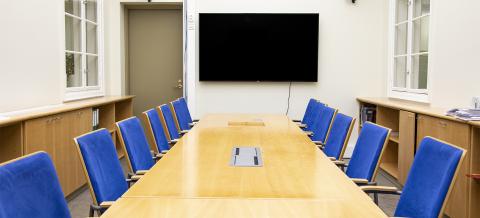 Pitkulaisessa tilassa on koko tilan täyttävä, vaaleaa puuta oleva pöytä, jota ympäröi siniset selkänojalliset tuolit. Takaseinällä on iso televisiä ja ovi. 