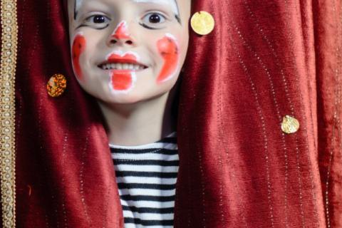 Lapsi kurkkaa verhon takaa ja hymyilee. Hänelle on maalattu pellen kasvot.