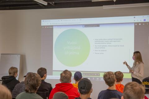 Opiskelijat katsovat, kun opettaja puhuu edessä ja näyttää valkokankaalta dioja.