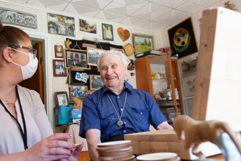 Ikääntynyt sinipaitainen mies istuu ja hymyilee nuoremman tummatukkaisen naisen vierellä.