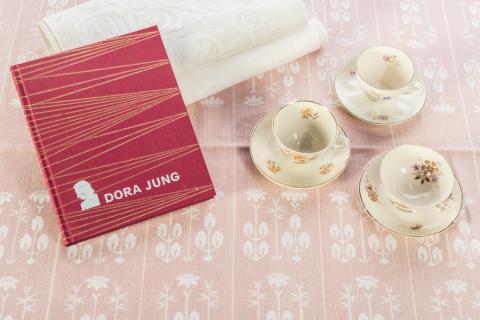 Kolme vaaleaa, vanhaa kahvikuppia on aseteltu vaaleanpunaiselle pellavapöytäliinalle punakantisen kirjan viereen.