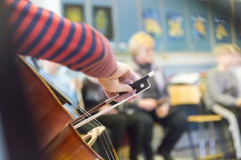 Lähikuva sellistin kädestä ja jousesta. Oppilaat kuuntelevat taustalla.