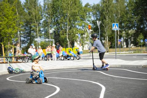 Liikennepuistossa pieni lapsi ajaa leikkimopolla ja isompi lapsi potkulaudalla.