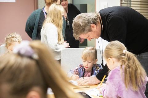 Lapsia ja aikuisia on keskittynyt tehtävään. Keskellä isä kumartuneena oman lapsensa työssä auttamiseen.