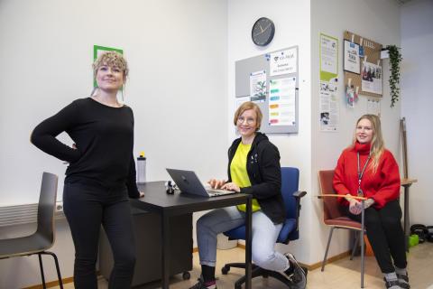 Kaksi henkilöä istuu ja kolmas seisoo lukion luokassa katsoen kameraan.