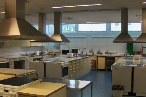Opetuskeittiöluokka, jossa on monta työskentelypistettä ja liesituulettimia katossa.