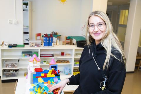 Smiling woman with eyeglasses looking at the camera. In front of her a table with toys.la hyllyillä lisää leluja.