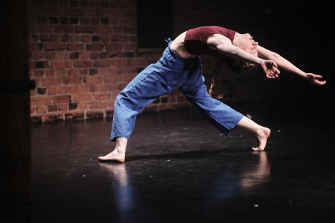 Tanssija taaksetaivutuksessa tiiliseinän edessä, päällä punainen toppi ja siniset housut.