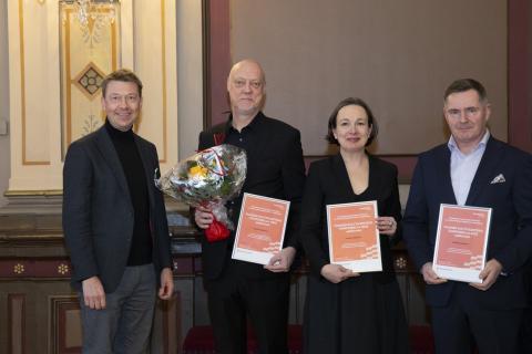 Four people in the row, three men and a woman. Woman and two men hold a diploma, one man also the bucket of flowers.