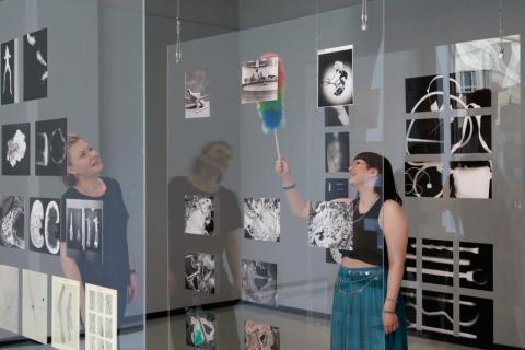 Kaksi naista ovat liiketilan sisällä. Toinen seuraa, kun toinen pyyhkii pölyjä pölyhuiskalla valokuvainstallaatiosta.