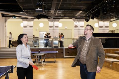 Rehtori ja apulaisrehtori testaavat koulun pingispeliä mailat käsissään. Pallo on korkealla ilmassa.