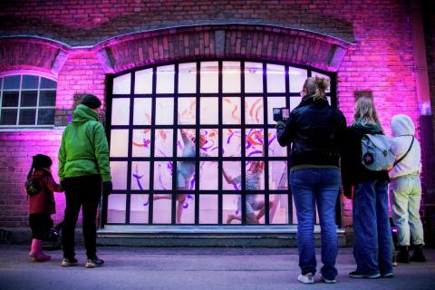 Kaksi taiteilijaa esiintyy ruutuikkunan takana ja eri ikäisiä ihmisiä seisoo ikkunan edessä katsomassa esitystä.