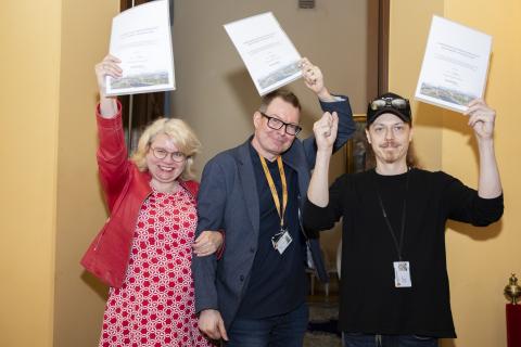 Kolme henkilöä rivissä heiluttelee päidensä yläpuolella papereita.