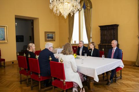 Kuusi ihmistä istuu valkoisella liinalla katetun pöydän ääressä. Kolme heistä on selin kameraan..