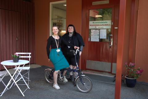  Mervi Matswetu sits on a folding bike and Emma Bragge stands beside her in front of Mansikkapaikka&#039;s red building.