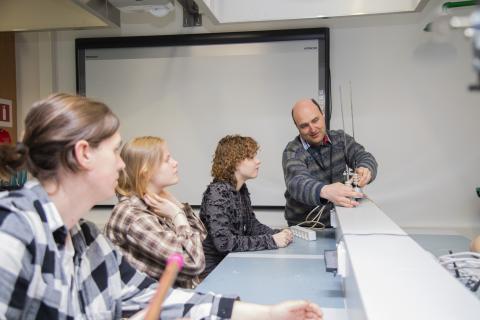 Kolme lukiolaista katselee, kun opettaja näyttää heille, miten pöydällä oleva tekninen laite toimii.