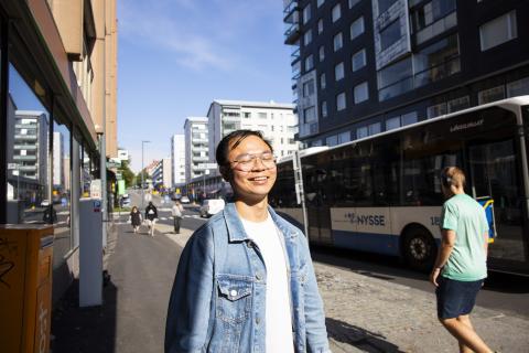 A person in a blue jacket standing in a sunny street, smiling.